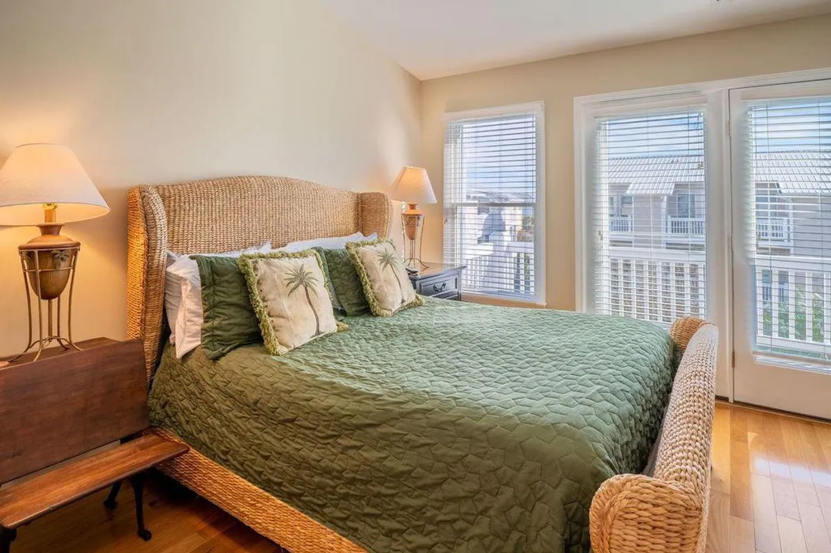 A serene bedroom featuring a woven bed frame topped with green bedding, palm-patterned pillows, and large windows that flood the room with natural light. The decor is tranquil and inviting, perfect for relaxing after a busy day.