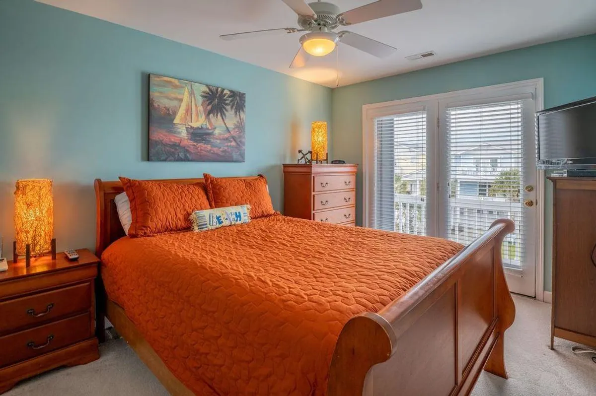 A cozy bedroom featuring a bed with vibrant orange bedding, palm tree artwork, wooden furniture, and lamps. The balcony view hints at the outdoors, creating a sense of connection to the surrounding area.