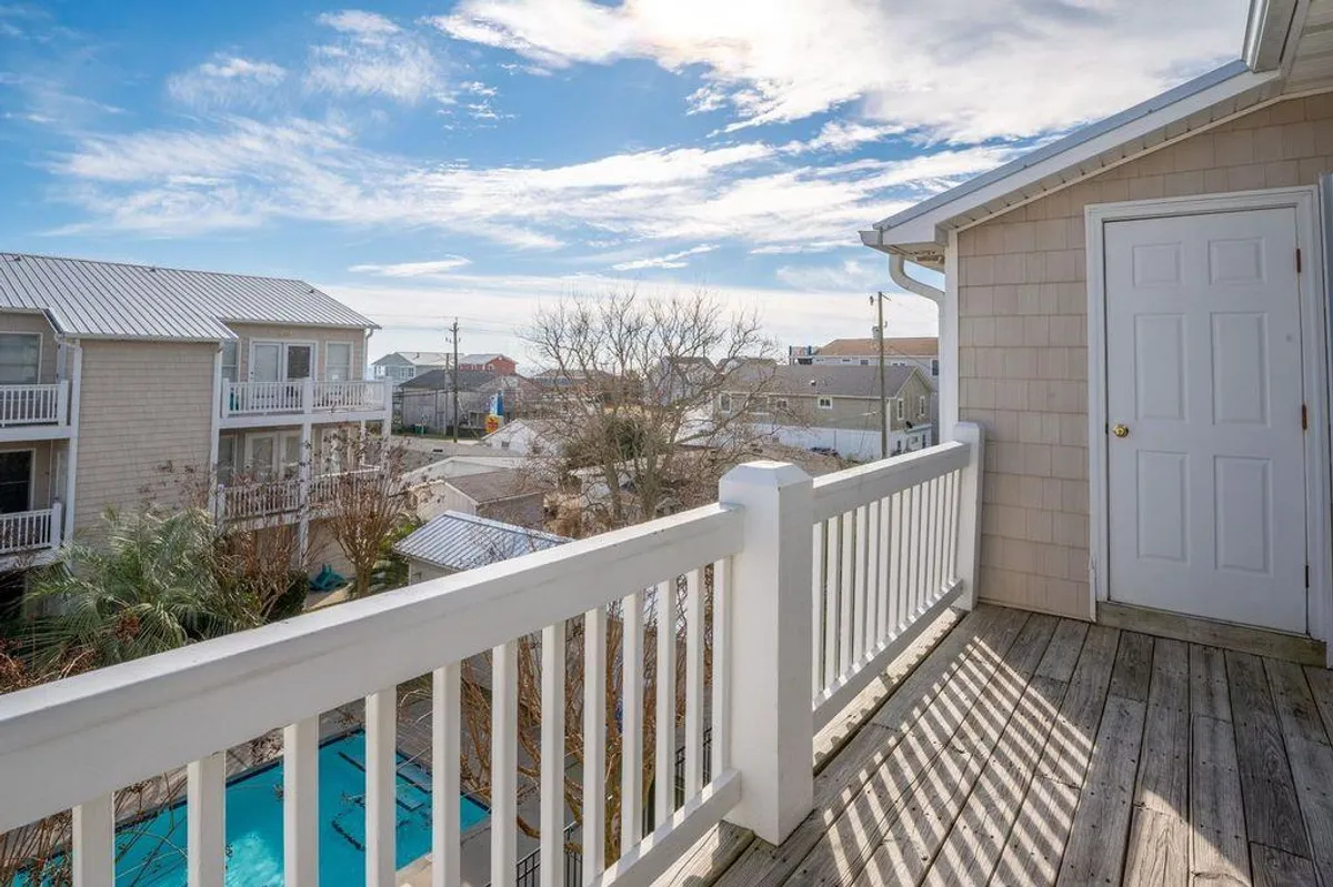 A peaceful balcony scene with railings overlooking the pool area, neighboring houses, and a bright blue sky. The view connects indoor comfort with outdoor amenities and community atmosphere.