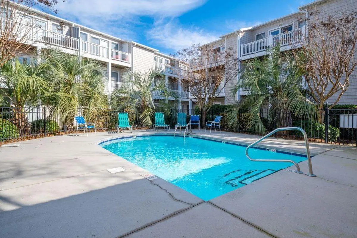 This exterior image captures a sparkling community swimming pool surrounded by lounge chairs, palm trees, and balconies. Sunlight reflects off the water, creating an inviting atmosphere for relaxation and recreation. The pool area is accented by greenery, offering a resort-like setting just steps from the residence.