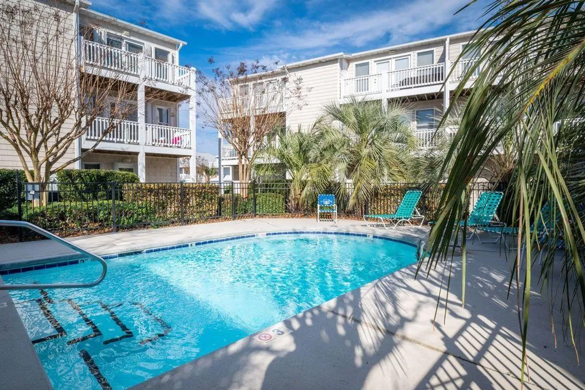 The image showcases the sun-drenched exterior of a spacious family home in Kure Beach, North Carolina. A sparkling swimming pool sits at the center, surrounded by inviting lounge chairs and swaying palm trees. The balconies of the house overlook this vibrant pool area, suggesting ample spaces for relaxation, socializing, and enjoying the coastal breeze. The scene exudes a laid-back, resort-style ambiance ideal for families and groups seeking fun and connection.
