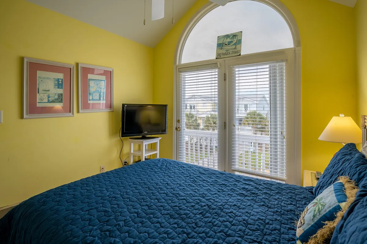 A vibrant living area with a blue quilted bed, large window, TV on a stand, and framed artwork. The room receives ample natural light, making it an inviting common area for relaxing, watching movies, or friendly group activities.