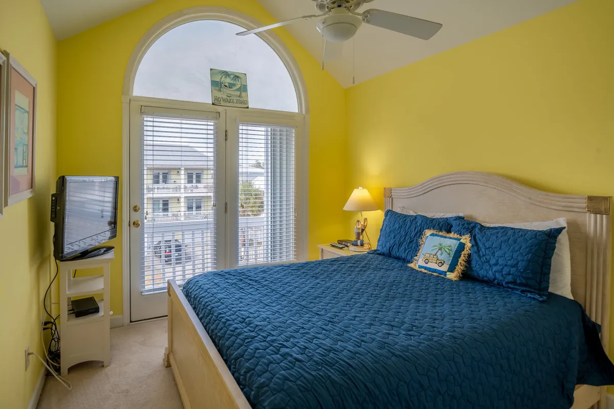 A bright, cheerful bedroom with yellow walls, a blue bedspread, large windows streaming in natural light, and a TV on a stand. The setting feels inviting, perfect for waking up to a sunny day and planning a day of festivities.