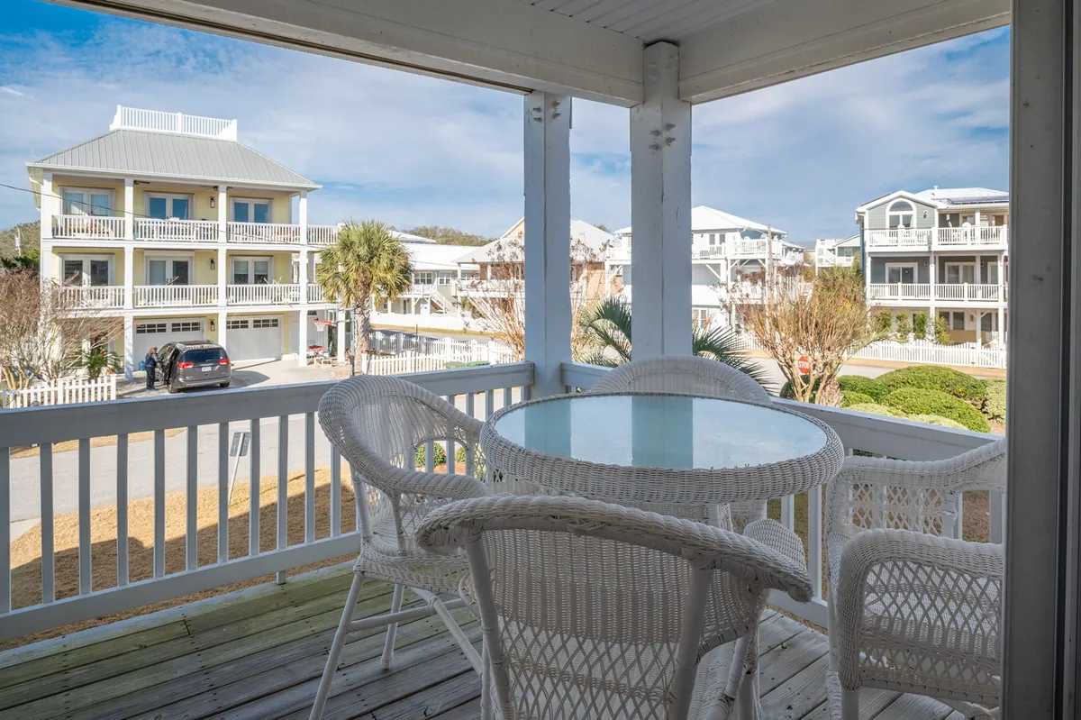 A relaxed outdoor deck setup featuring wicker furniture and a glass table, with views of neighboring houses. The space is clearly geared toward enjoying meals, morning coffee, or sunset drinks in the fresh air.