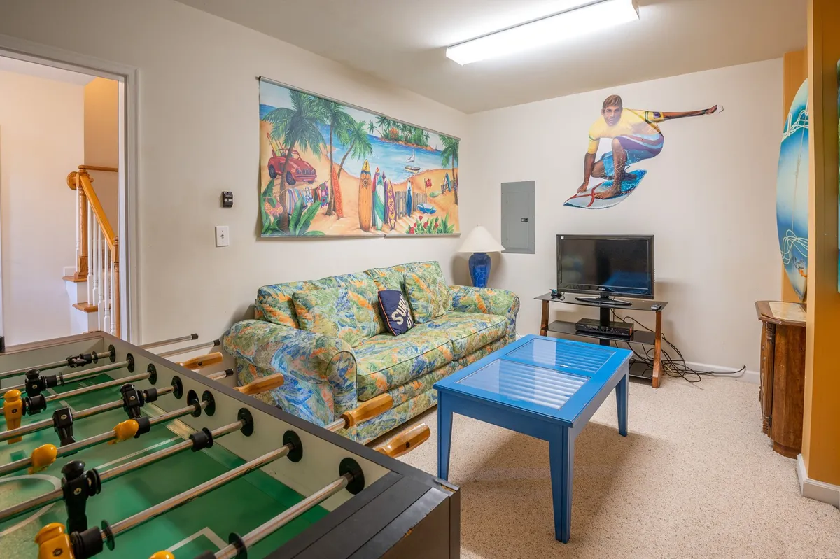 A cozy yet lively basement setup highlighted by a colorful couch, foosball table, blue coffee table, and a bold surfing mural. The scene invites group activities like foosball tournaments, watching TV, or relaxing on the sleeper sofa, all in a relaxed, inviting environment.