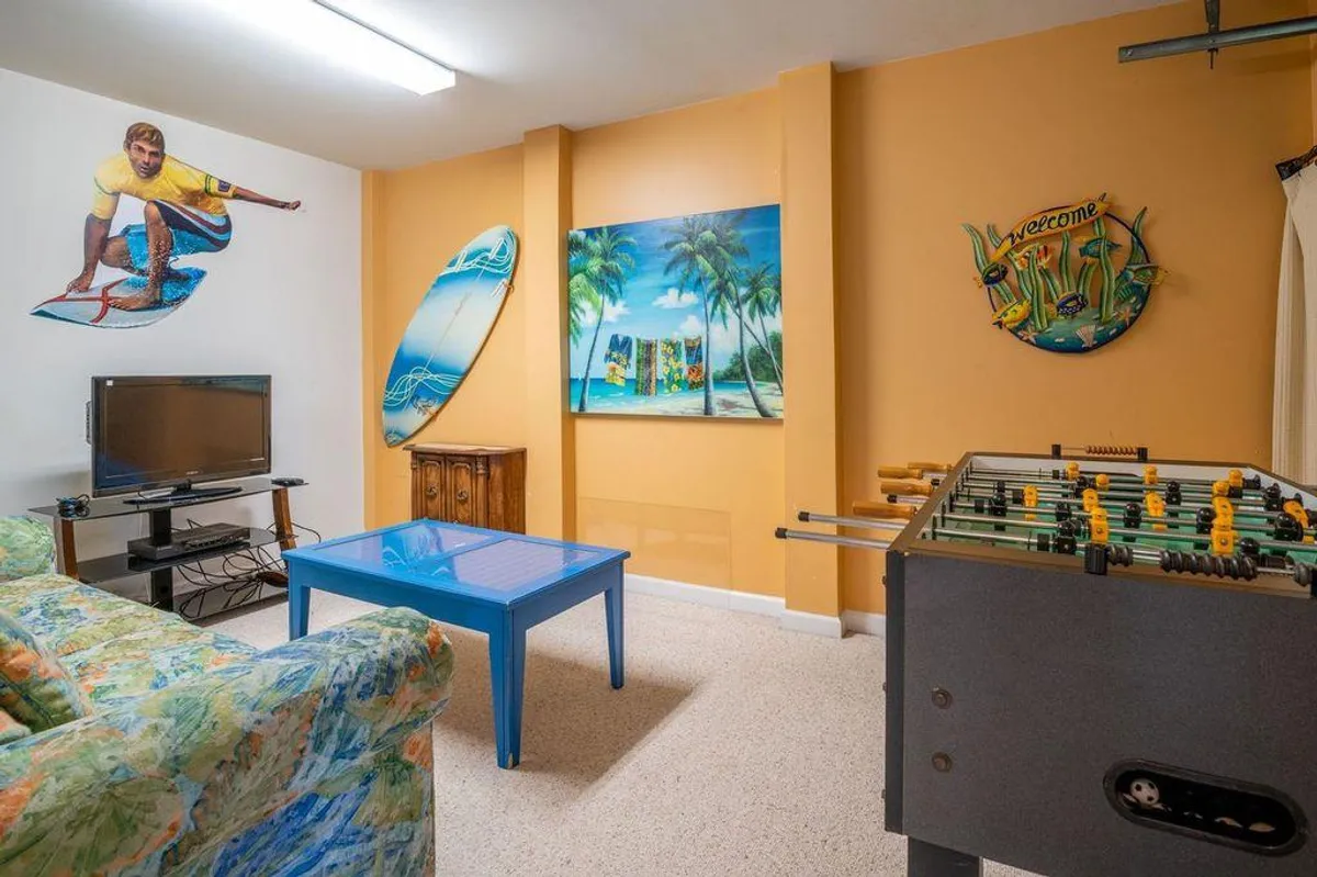This vibrant basement scene features a colorful couch and foosball table set against a striking surfer mural and ocean-themed artwork. The arrangement captures a playful, laid-back atmosphere with clear zones for both games and relaxation. Natural light streams in, and the proximity to the blue coffee table and TV indicates spaces for movie nights and social gatherings.