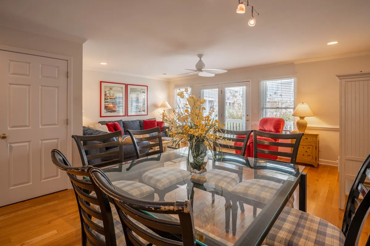 A welcoming dining area with a glass dining table, a cheerful yellow floral centerpiece, colorful red and blue accent chairs, and artistic decor on the walls. The space radiates warmth and is ideal for shared meals or board game nights.