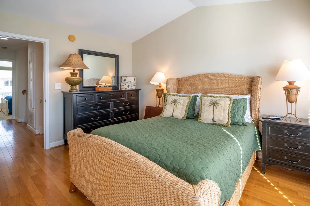 A calm, cozy bedroom with a woven bed frame, green quilt, decorative pillows with palm trees, and matching nightstands with soft lamp lighting. The space feels soothing and designed for comfort.