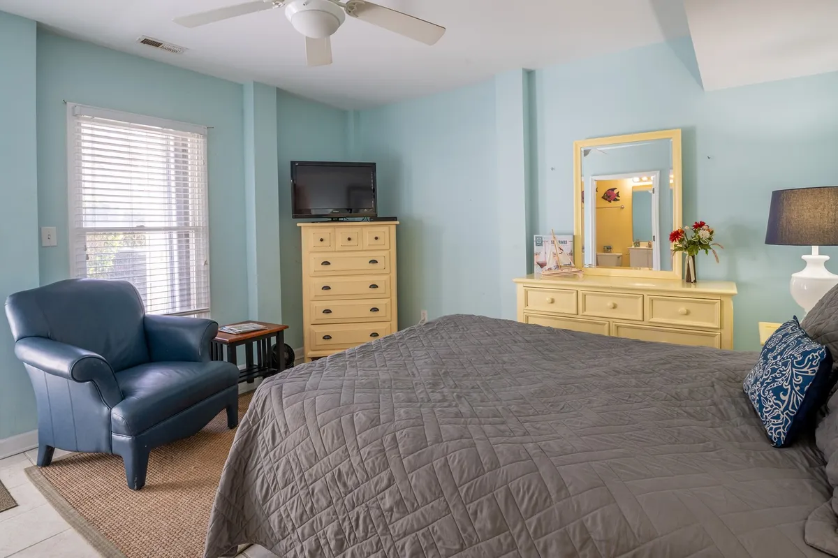 A tranquil bedroom scene with a bed featuring a gray quilt, a yellow dresser, a blue armchair, and a decorative lamp. Large windows illuminate the space with bright sunlight, creating an inviting spot for leisurely mornings or quiet reading, just steps away from a balcony setting.