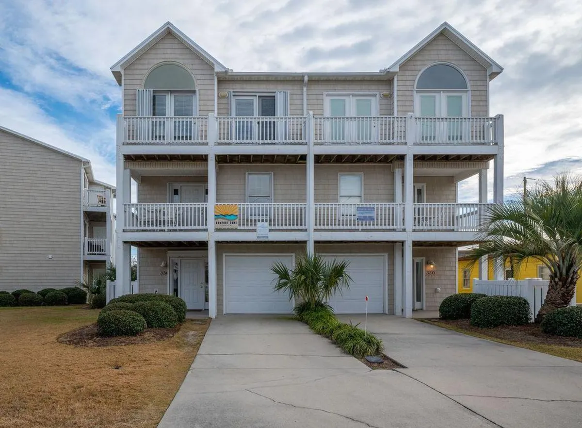 The image shows the multi-level home's exterior, with balconies shaded by palm trees and an inviting entryway below. The balconies offer views toward the coastline, while the surrounding yard hints at outdoor gatherings and easy access to the beach beyond.