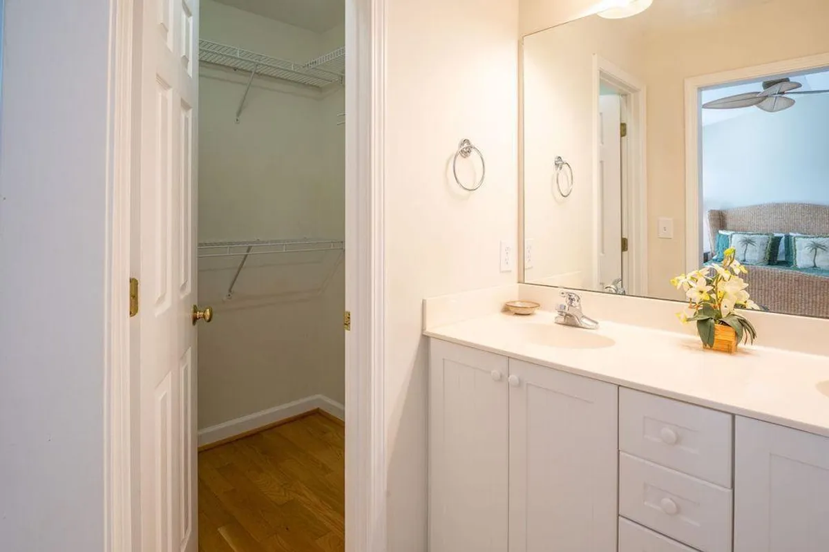 A bright and organized bathroom featuring a vanity with sink, large mirror, flowers adding a pop of color, and a closet door nearby. The ambiance is clean and spa-like, ideal for morning routines or winding down.